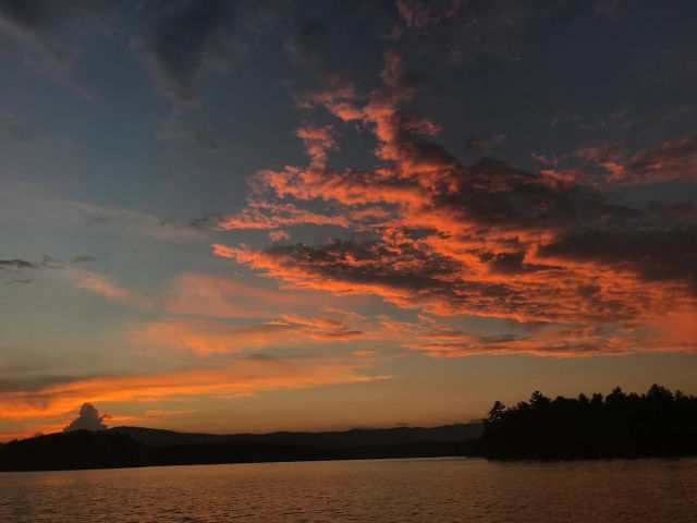 Fiery orange sunset over a calm lake, dramatic glowing clouds streaking the evening sky above a dark silhouetted treeline and distant hills.