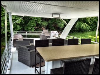 Covered waterfront deck with wicker lounge chairs, bar counter and grill overlooking lush green shoreline.