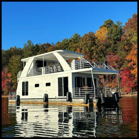 Modern white houseboat floating on a calm lake with vibrant fall foliage on the shore, clear blue sky and crisp water reflections.