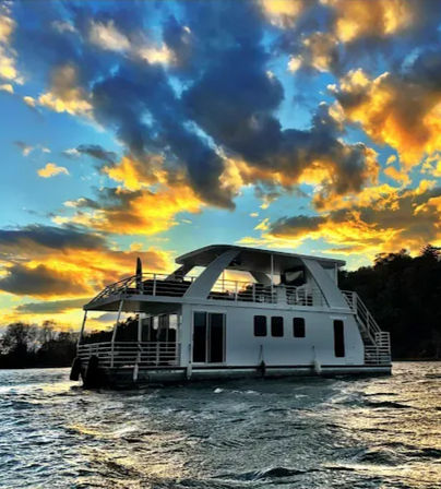 White modern houseboat cruising on a lake at sunset with dramatic orange-and-blue clouds, choppy water, and a tree-lined shoreline.