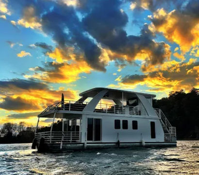 White modern houseboat cruising on a lake at sunset with dramatic orange-and-blue clouds, choppy water, and a tree-lined shoreline.
