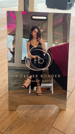 Polished mirror display with circular logo for a permanent-jewelry service reflecting a woman in a black dress and strappy black heels taking a mirror selfie in a bright indoor lounge with wooden floors and pink accent lighting.