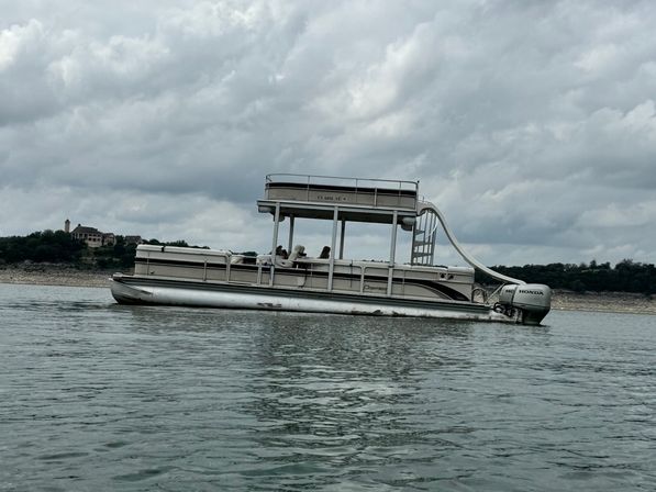 Two-level pontoon party boat with a waterslide and Honda outboard floating on a calm lake under a moody cloudy sky, rocky shoreline and a distant lakeside house in the background.