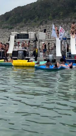 Summer lake party with people on connected pontoon boats featuring twin water slides, colorful inflatables and paddleboards floating near a rocky shoreline and tree-covered hill.