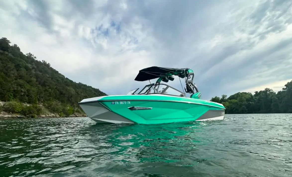 Teal wakeboard boat with black wake tower and folded bimini floating on a green lake near forested hills under a dramatic cloudy sky