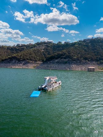 Pontoon boat with a waterslide and blue floating mat on a green lake, rocky hillside with houses under a bright blue sky and puffy clouds.