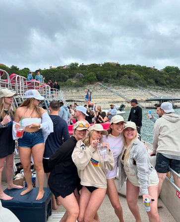 Crowd of young adults posing and laughing on a crowded pontoon boat at a lakeside party, wearing caps and casual summer outfits and holding drinks, with rocky shoreline and overcast sky in the background.