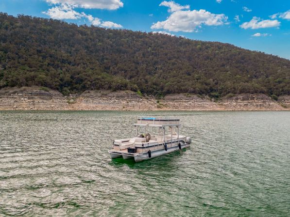 Leisure pontoon boat floating on a green lake near a rocky shoreline with forested hills and a blue sky dotted with clouds