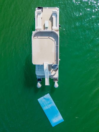 Aerial drone view of a white pontoon boat with a rear water slide, twin outboard motors and a blue floating mat drifting on clear green lake water.