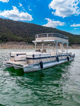 Double-deck pontoon boat floating on green lake water near a rocky shoreline and wooded hills under a sunny blue sky with fluffy clouds