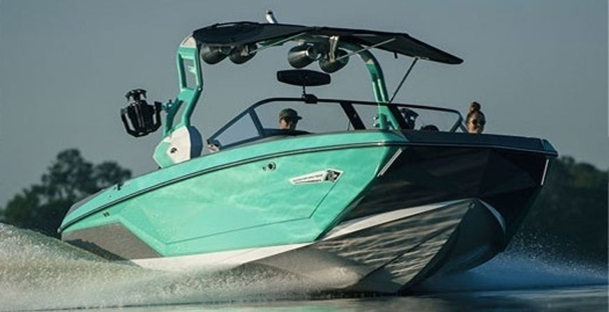Teal wakeboard boat with tower and speakers skimming a lake, kicking up spray and wake with two passengers and a tree-lined shore in the background.