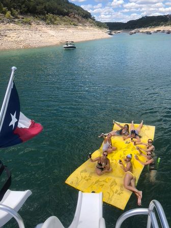 Friends lounging on a bright yellow float in a Texas lake, toasting drinks near a boat slide with a Texas flag and rocky shoreline under blue skies.