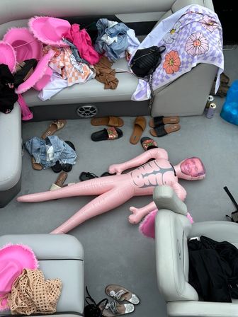 Pontoon boat interior strewn with sandals, denim, colorful towels and pink feathered cowboy hats, plus a large inflatable pink doll with a smiling printed face.
