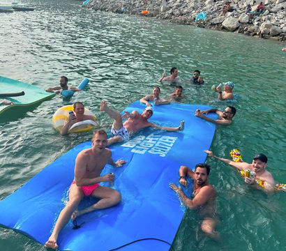 Group of friends relaxing on and around a large blue inflatable water mat in clear green water near a rocky shoreline, enjoying drinks on a sunny summer day.