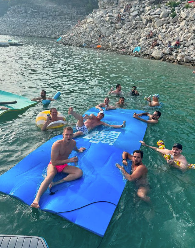 Group of friends relaxing on and around a large blue inflatable water mat in clear green water near a rocky shoreline, enjoying drinks on a sunny summer day.