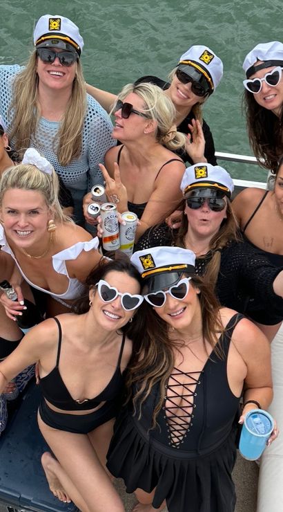 Group of women in swimsuits and captain hats smiling on a boat deck over green water, many wearing sunglasses (including heart-shaped), holding canned drinks and cups, posing for a lively summer boat party photo.