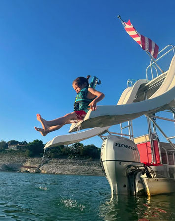 Child in a blue life jacket launching off a boat waterslide into a lake, water splashing below under a clear blue sky with an American flag flying from the boat.