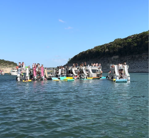 Summer lake party: cluster of houseboats with white water slides and colorful kayaks and paddleboards anchored near a rocky, tree-lined shoreline under a clear blue sky