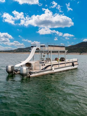 Double-decker pontoon boat with waterslide and twin outboard motors floating on a lake near a hilly shoreline under a bright blue, partly cloudy sky.