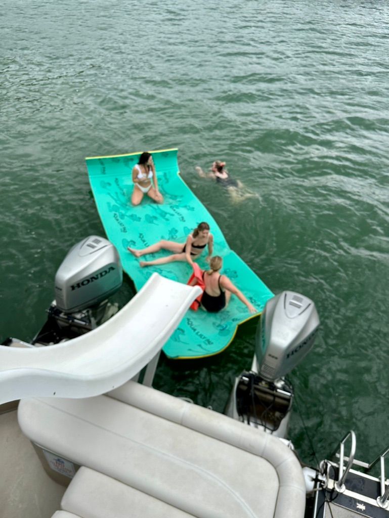 Group lounging on a bright turquoise floating mat next to a pontoon boat with twin Honda outboard motors and slide on a green lake