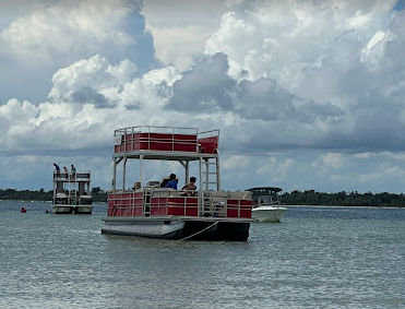 Red double-decker pontoon boat anchored in calm bay waters under dramatic cumulus clouds, with another pontoon and a small motorboat near a tree-lined shoreline.
