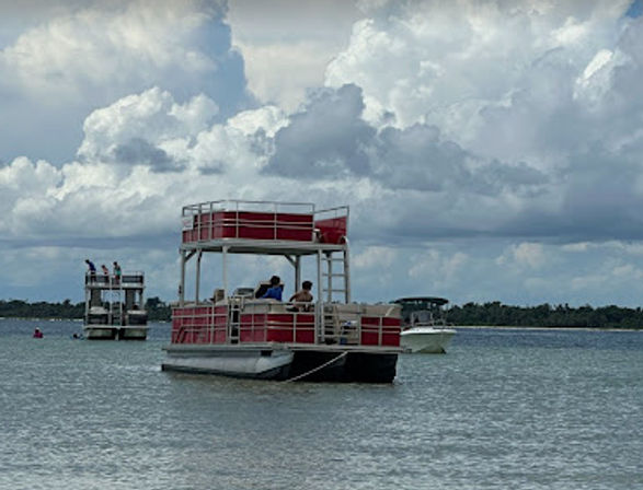 Red double-decker pontoon boat anchored in calm bay waters under dramatic cumulus clouds, with another pontoon and a small motorboat near a tree-lined shoreline.