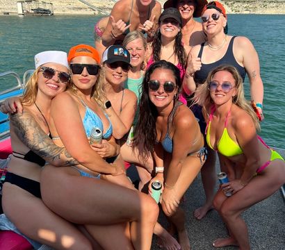 Group of smiling women in colorful bikinis posing on a boat, holding canned drinks at a sunny lake party by a rocky shoreline and wooded hillside.