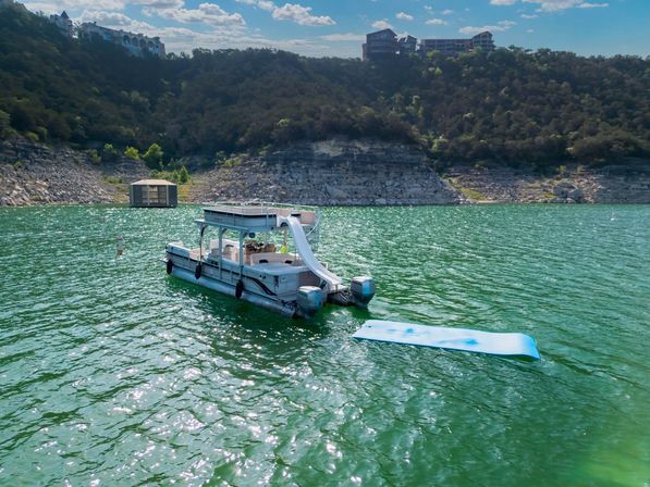 Pontoon boat with white waterslide and blue floating mat on an emerald-green lake, rocky cliff shoreline and hillside homes in the background under a sunny blue sky.