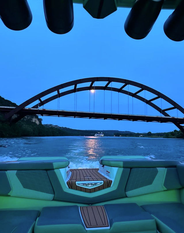View from the stern of a boat with mint-green seats and teak deck cruising beneath a large arched steel bridge at blue twilight, moonlight reflecting on the river and the boat’s wake trailing toward the distant shoreline.