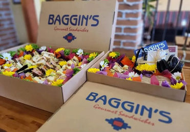 Open catering boxes of assorted finger sandwiches with colorful tissue paper, daisies, and utensils on a wooden table.