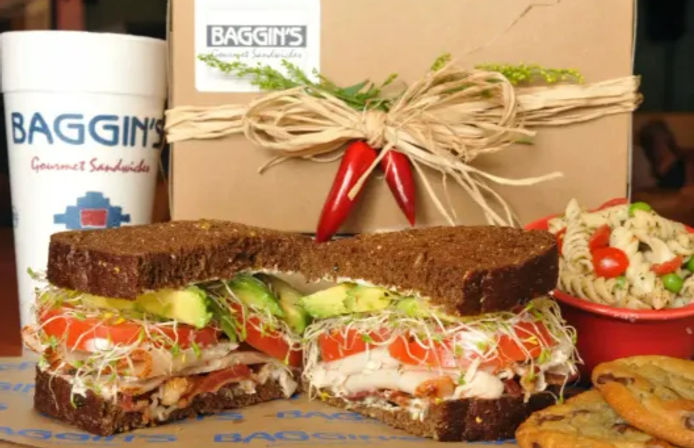 Gourmet deli-style turkey and avocado sandwich on dark whole-grain bread with sprouts, tomato and bacon, shown with a pasta salad side, cookies and a to-go drink and boxed takeout.