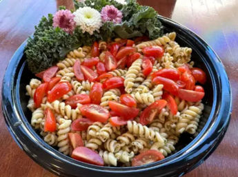 Bright herbed rotini pasta salad tossed with cherry tomato halves in a black bowl, garnished with curly kale and flowers on a wooden table.
