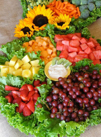 Vibrant summer fruit and veggie platter with red grapes, strawberries, cubed watermelon, cantaloupe and pineapple, kiwi slices around a creamy dip, nestled on green lettuce with broccoli, baby carrots and decorative sunflowers.
