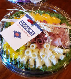 Round clear plastic catering platter with rolled provolone, deli ham and cheddar slices, lettuce garnish and raffia-tied decorative tag on a wooden table