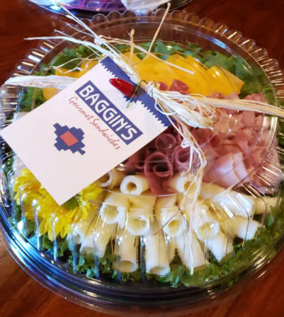 Clear plastic party platter of rolled deli meats and cheese slices on a bed of lettuce with yellow pepper strips, tied raffia bow and small gift tag — catered deli tray