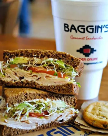 Stacked turkey and cheese sandwich on rye with tomato, sliced avocado and alfalfa sprouts, paired with a cookie and a takeout drink cup on a wooden deli table.