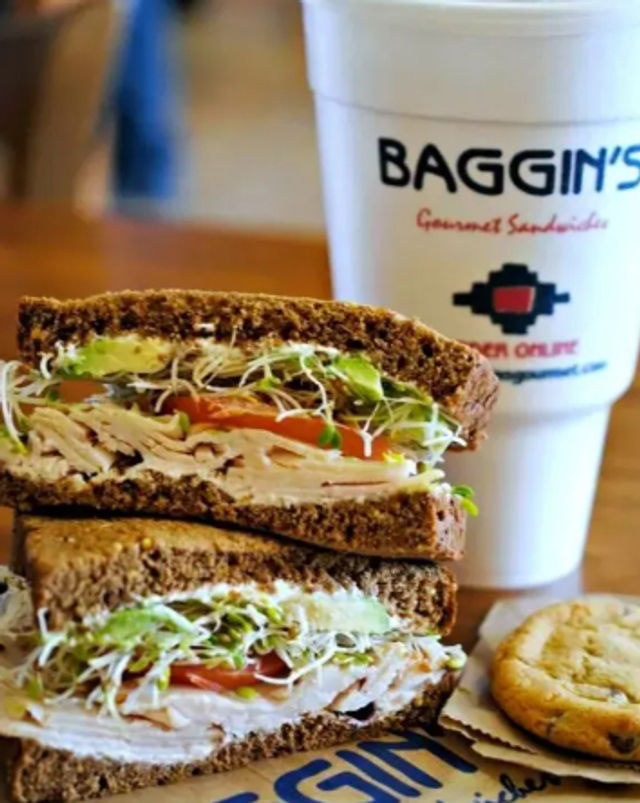 Stacked turkey and cheese sandwich on rye with tomato, sliced avocado and alfalfa sprouts, paired with a cookie and a takeout drink cup on a wooden deli table.