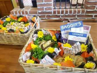Wicker catering baskets on a wooden table filled with assorted sandwiches, cookies, leafy greens, orange wrappers and bright yellow flowers, with catering brochures and a brick interior in the background.