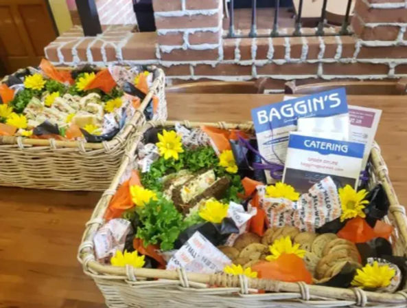 Two wicker catering baskets on a wooden table holding assorted deli sandwiches, wrapped sides, cookie assortment, leafy greens and bright yellow sunflower garnishes, with catering brochures and a brick interior backdrop.