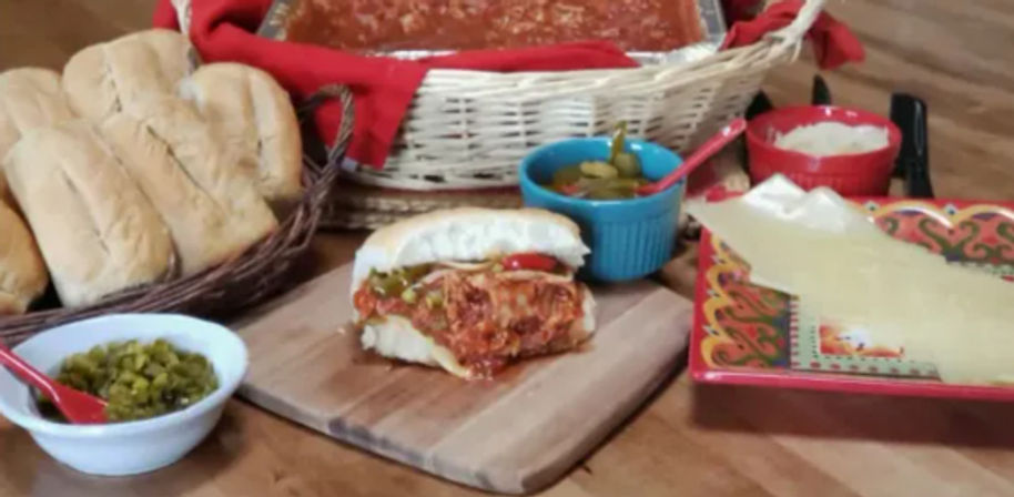 Deli-style Italian meatball sub on a wooden board with marinara sauce, crusty rolls in a basket, bowls of pickles and peppers, and grated cheese on a colorful plate.