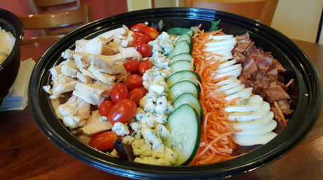 Colorful Cobb-style salad in a black takeout bowl on a wooden table, with rows of sliced grilled chicken, cherry tomatoes, blue cheese crumbles, cucumber rounds, shredded carrots, sliced hard-boiled egg and bacon.