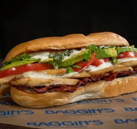 Close-up of a hearty sub sandwich on a toasted roll with sliced deli meat and crispy bacon, melted white cheese, tomato slices, fresh lettuce, avocado, and mayo.