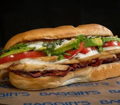 Close-up of a hearty sub sandwich on a toasted roll with sliced deli meat and crispy bacon, melted white cheese, tomato slices, fresh lettuce, avocado, and mayo.