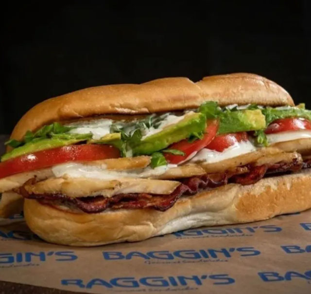 Close-up of a hearty sub sandwich on a toasted roll with sliced deli meat and crispy bacon, melted white cheese, tomato slices, fresh lettuce, avocado, and mayo.