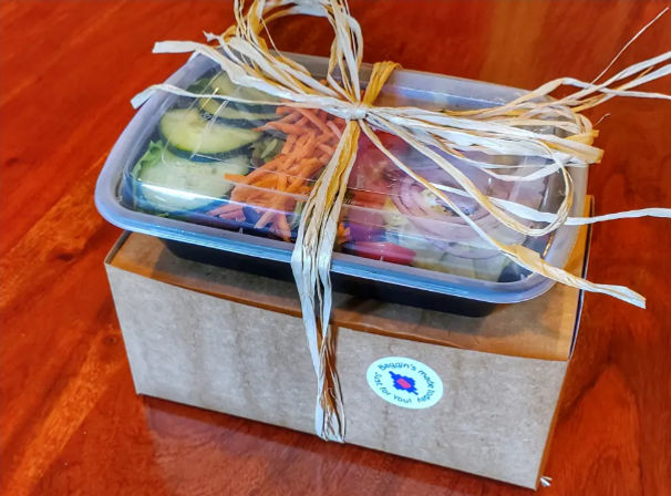 Colorful takeout salad with cucumber, shredded carrots, red onion and tomato in a clear container tied with a raffia bow on a brown cardboard box set on a wooden table