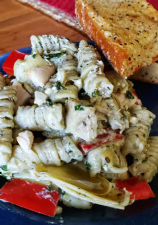 Creamy pesto rotini pasta salad with chicken, marinated artichokes, red bell pepper and herbs, served with garlic bread on a blue plate over a wooden table.