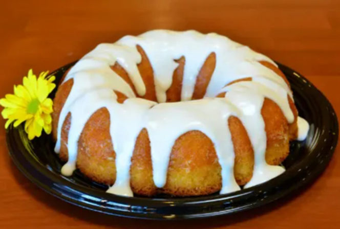 Glazed bundt cake with thick white icing dripping down the sides on a black serving plate, garnished with a bright yellow daisy on a wooden table.
