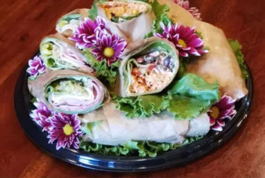 Assorted deli wrap platter with sliced pinwheel wraps filled with ham, turkey, chicken salad, lettuce and cheese, arranged on green leaf lettuce and garnished with purple daisy flowers on a black serving tray.
