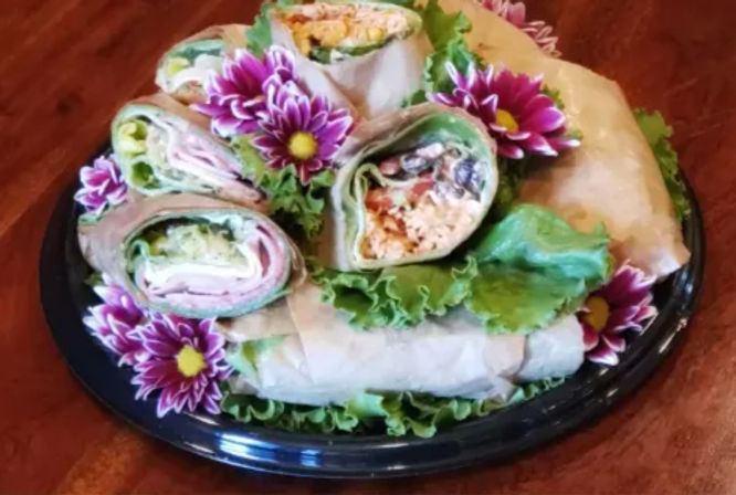 Colorful party platter of assorted deli sandwich wraps sliced into pinwheels, layered on lettuce and garnished with purple chrysanthemum flowers on a black tray.