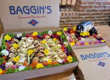 Colorful catering sandwich platter — two open cardboard boxes of gourmet mini sandwiches garnished with lettuce and bright edible flowers, arranged on a wooden table against a brick-wall café backdrop.